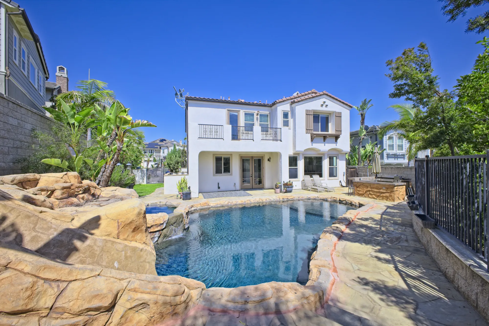 Backyard of a two-story white house with a rock-edged swimming pool, lounge chairs, and tropical plants under a clear blue sky.