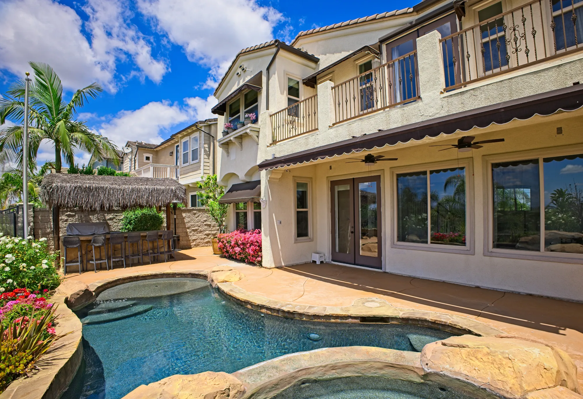Backyard of a two-story house featuring a swimming pool, hot tub, patio with ceiling fans, and a thatched tiki bar with bar stools.