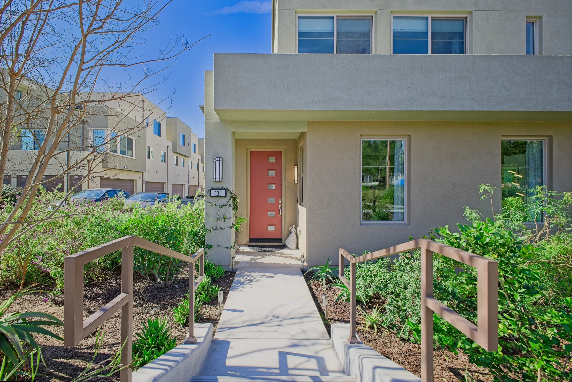 Modern beige townhouse entrance with a red front door, flanked by two windows and greenery along a concrete path with handrails.