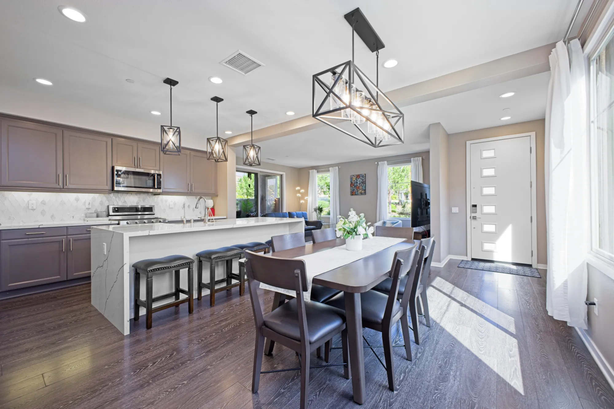 Modern open kitchen and dining area with dark wood floors, gray cabinets, large island with stools, and dining table with six chairs under geometric pendant lights.