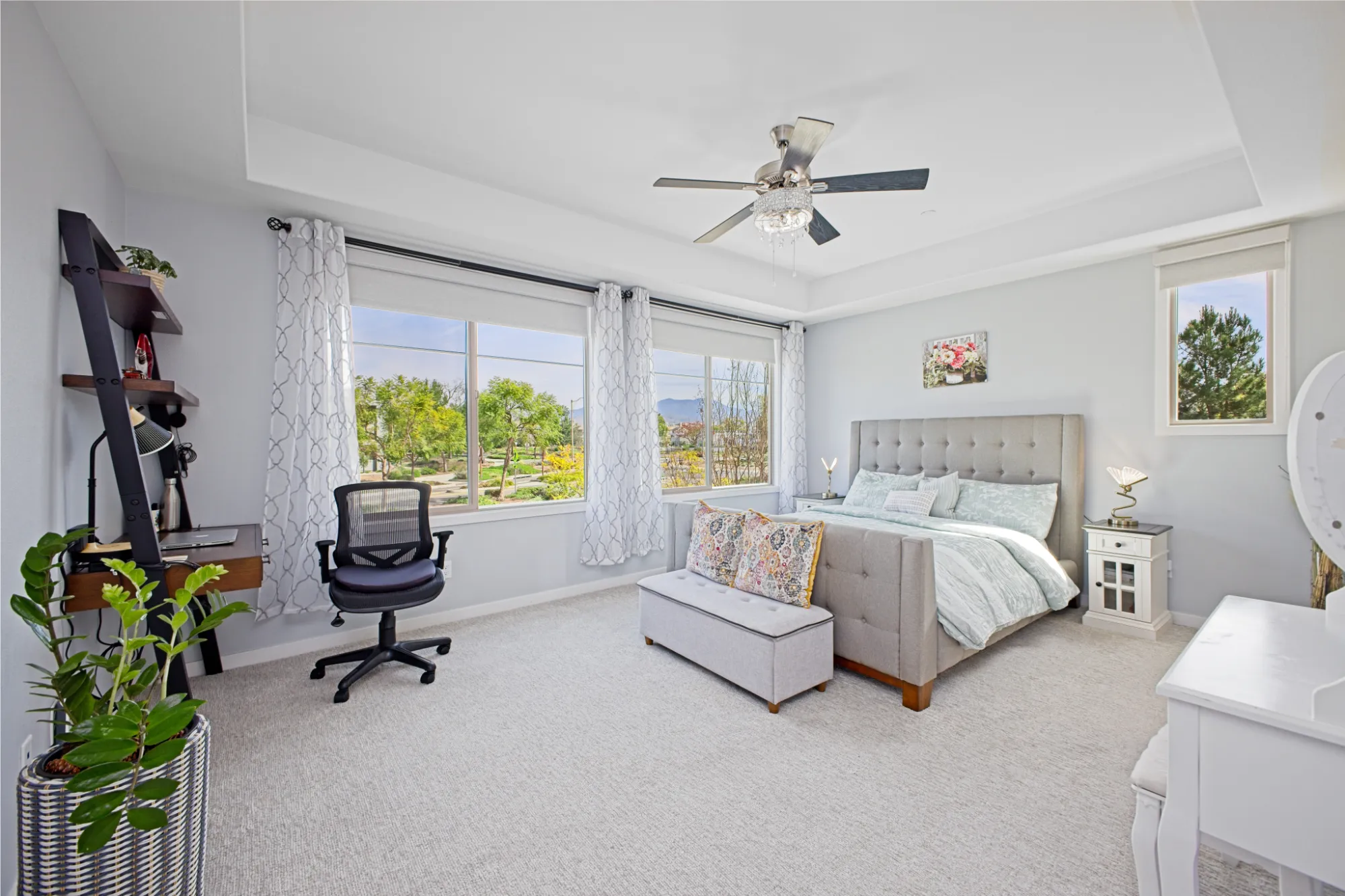 Bright bedroom with large windows, a gray upholstered bed with decorative pillows, a small bench at the foot of the bed, a black office chair, a wooden desk with shelves, and a ceiling fan.