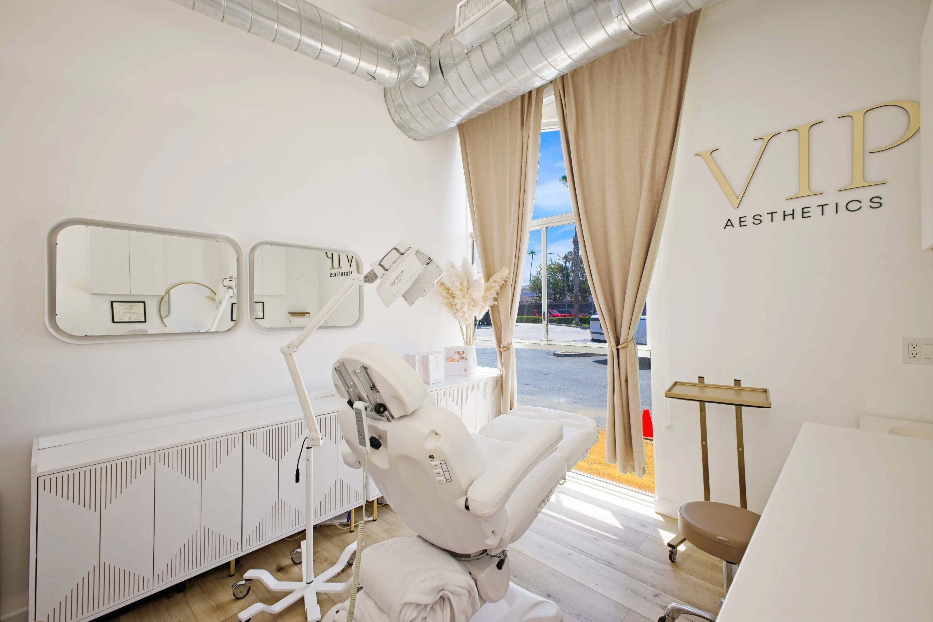Bright aesthetic clinic treatment room with a white adjustable chair, medical light, beige curtains, and VIP Aesthetics sign on the wall.
