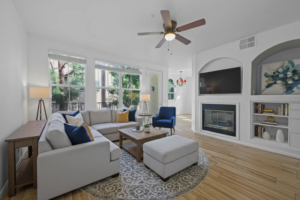 Bright living room with light gray sectional sofa, blue armchair, wooden coffee table, round patterned rug, ceiling fan, and built-in shelves with TV and fireplace.