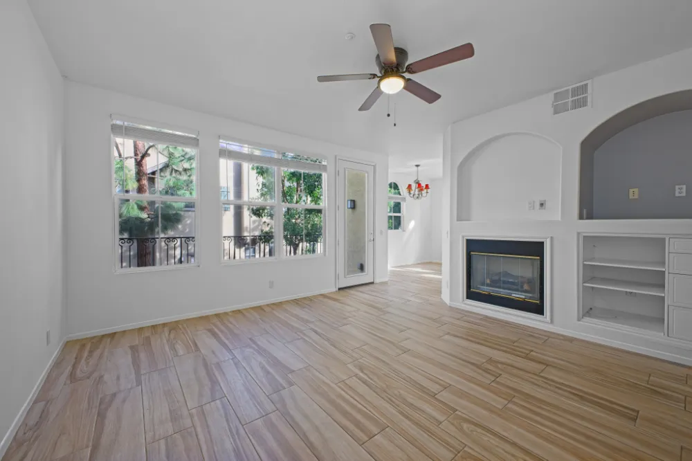 Empty living room with light wood flooring, white walls, a ceiling fan, large windows, a fireplace, and built-in shelving.