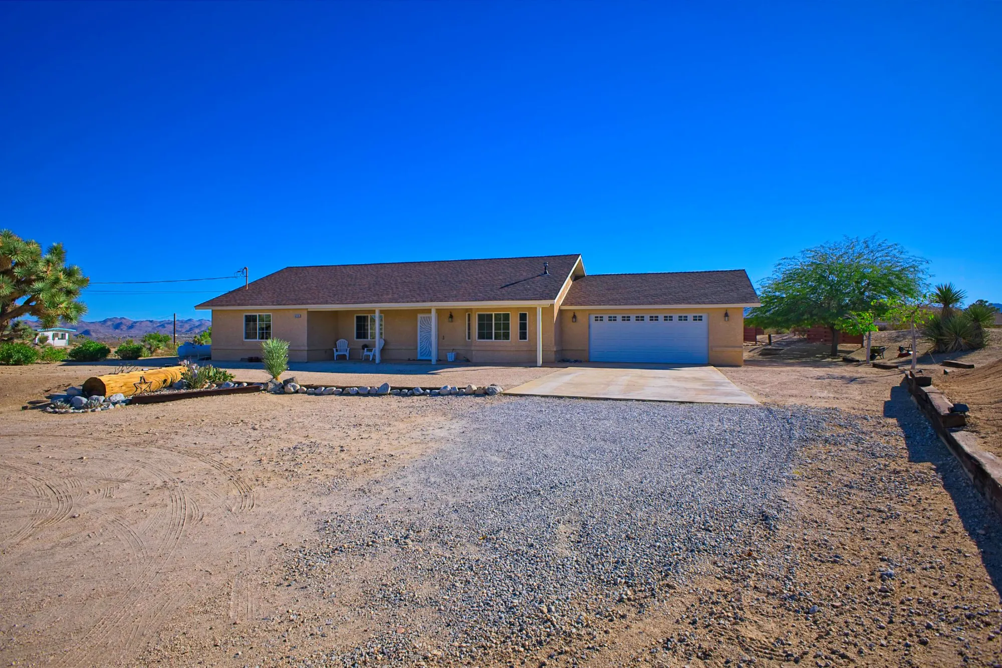 Single-story beige house with a brown roof, two white chairs on the front porch, and a double garage under a clear blue sky in a desert landscape.