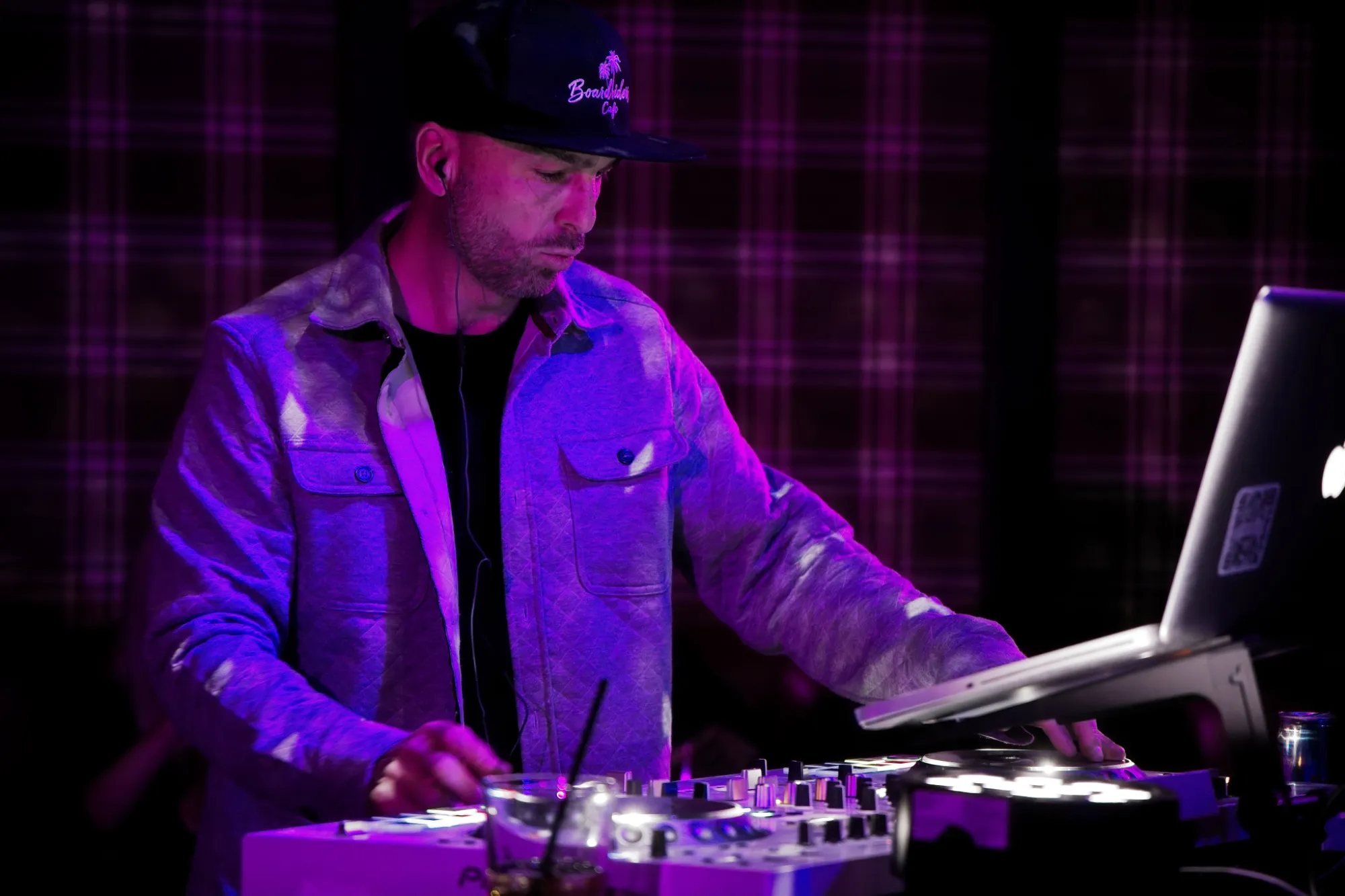 DJ wearing a cap and jacket mixing music using a controller and laptop under purple lighting.
