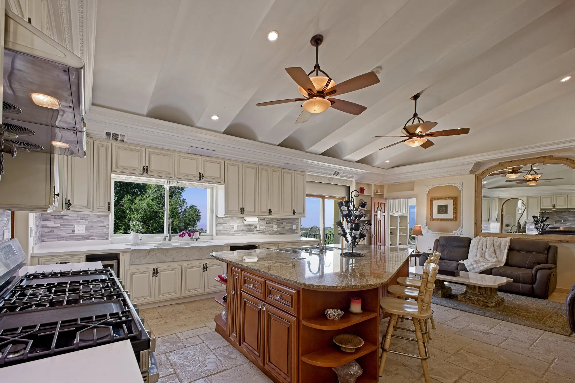 Spacious kitchen with large granite island, wooden stools, ceiling fans, and large window with outside view.