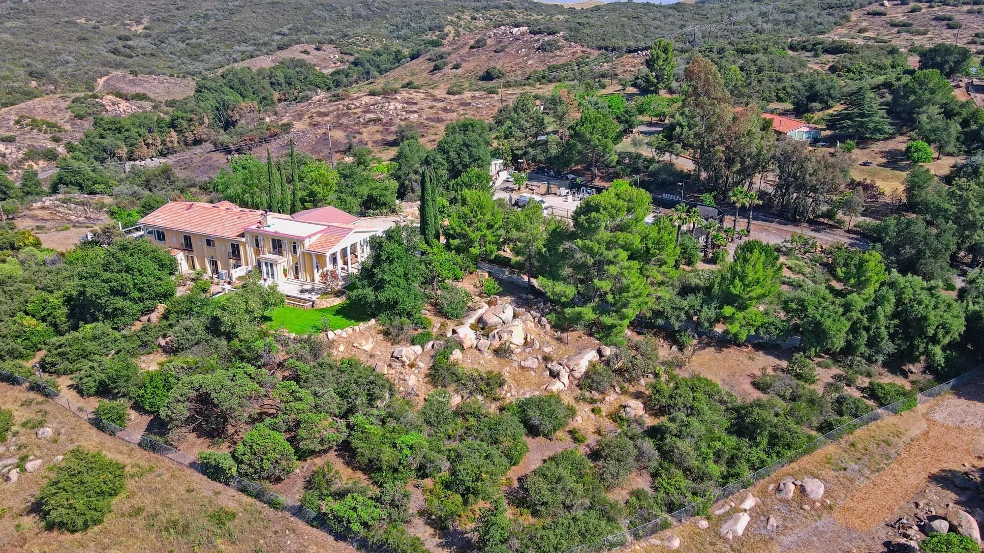 A large yellow house with a red tile roof surrounded by green trees and rocky terrain.