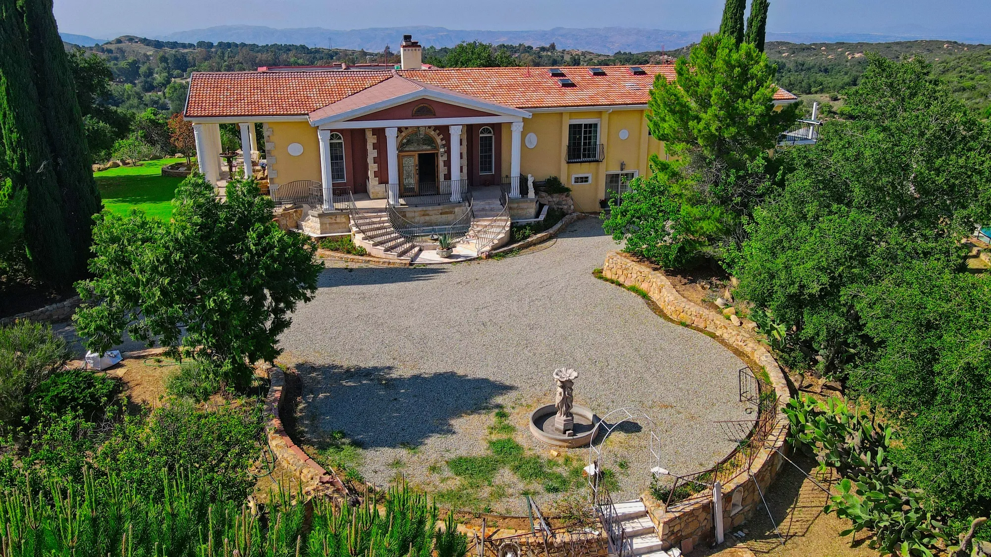 A large yellow house with a tiled roof, surrounded by green trees and a stone circular driveway.