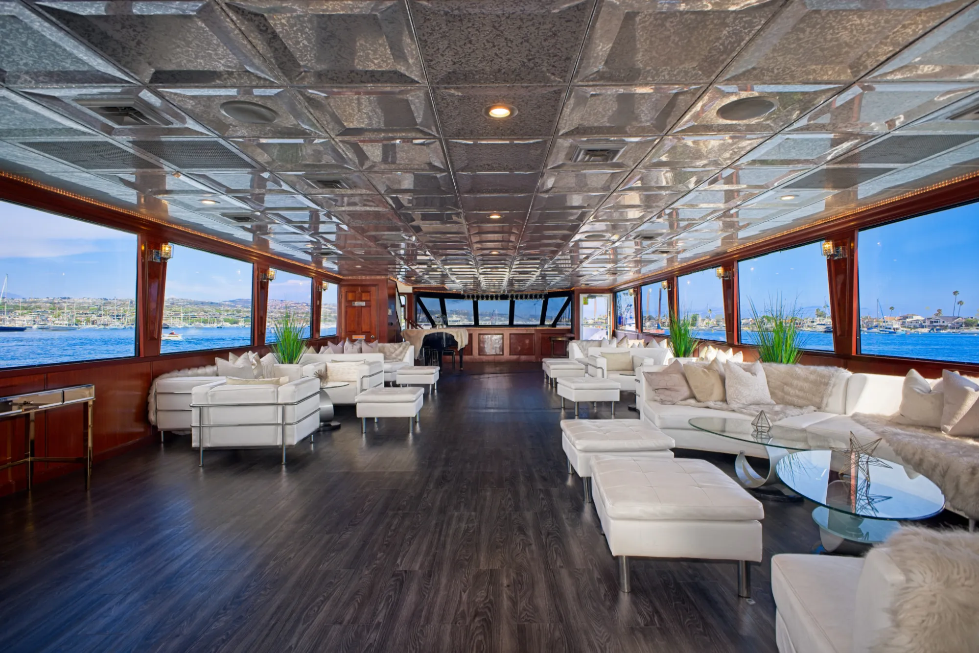 Spacious yacht lounge with white seating, glass tables, plants, and panoramic windows showing water views.