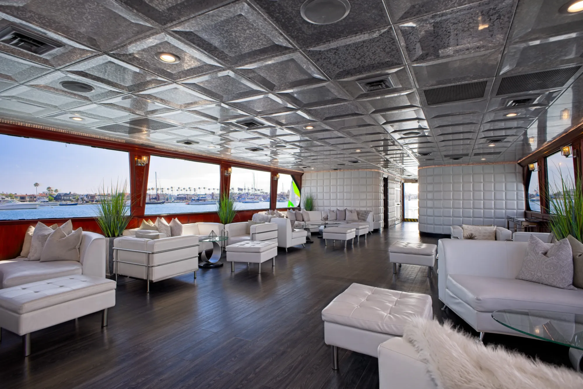 Luxurious yacht lounge with white sofas, large windows showing boats and calm water outside.