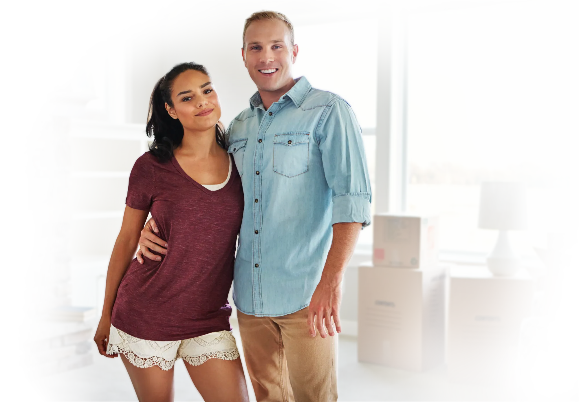 Smiling young couple standing in a room with moving boxes and a window behind them.