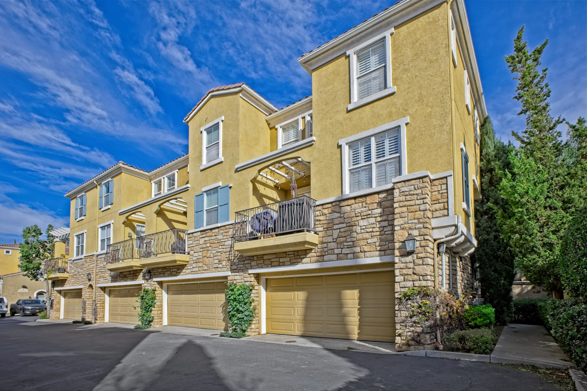 Yellow townhouse complex with stone accents, balconies, garages, and tall green trees under a blue sky.