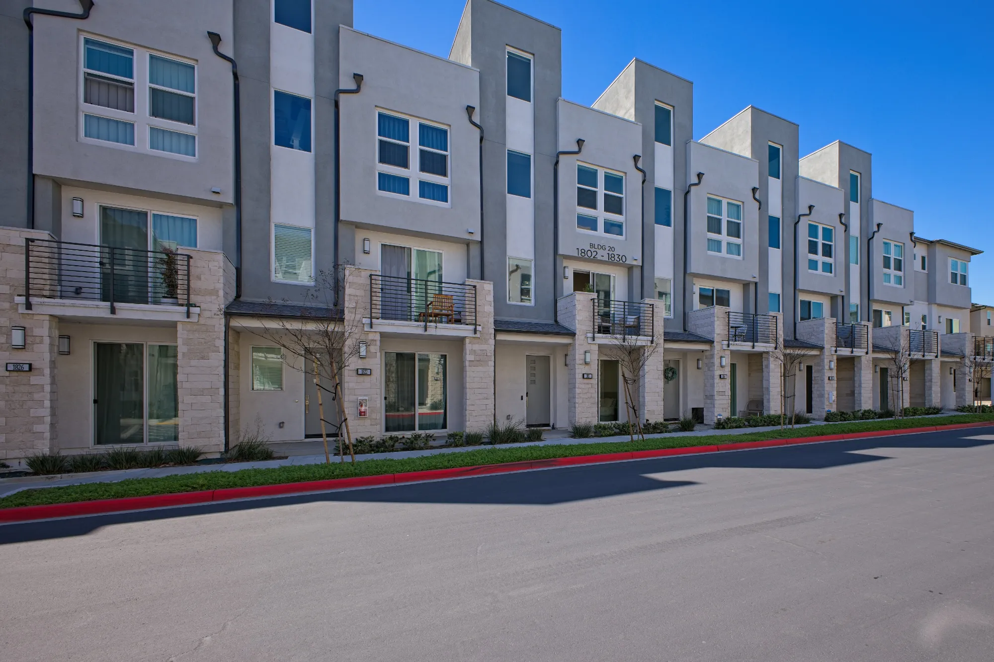 Row of modern gray townhouses with balconies, small trees, and clear blue sky above.