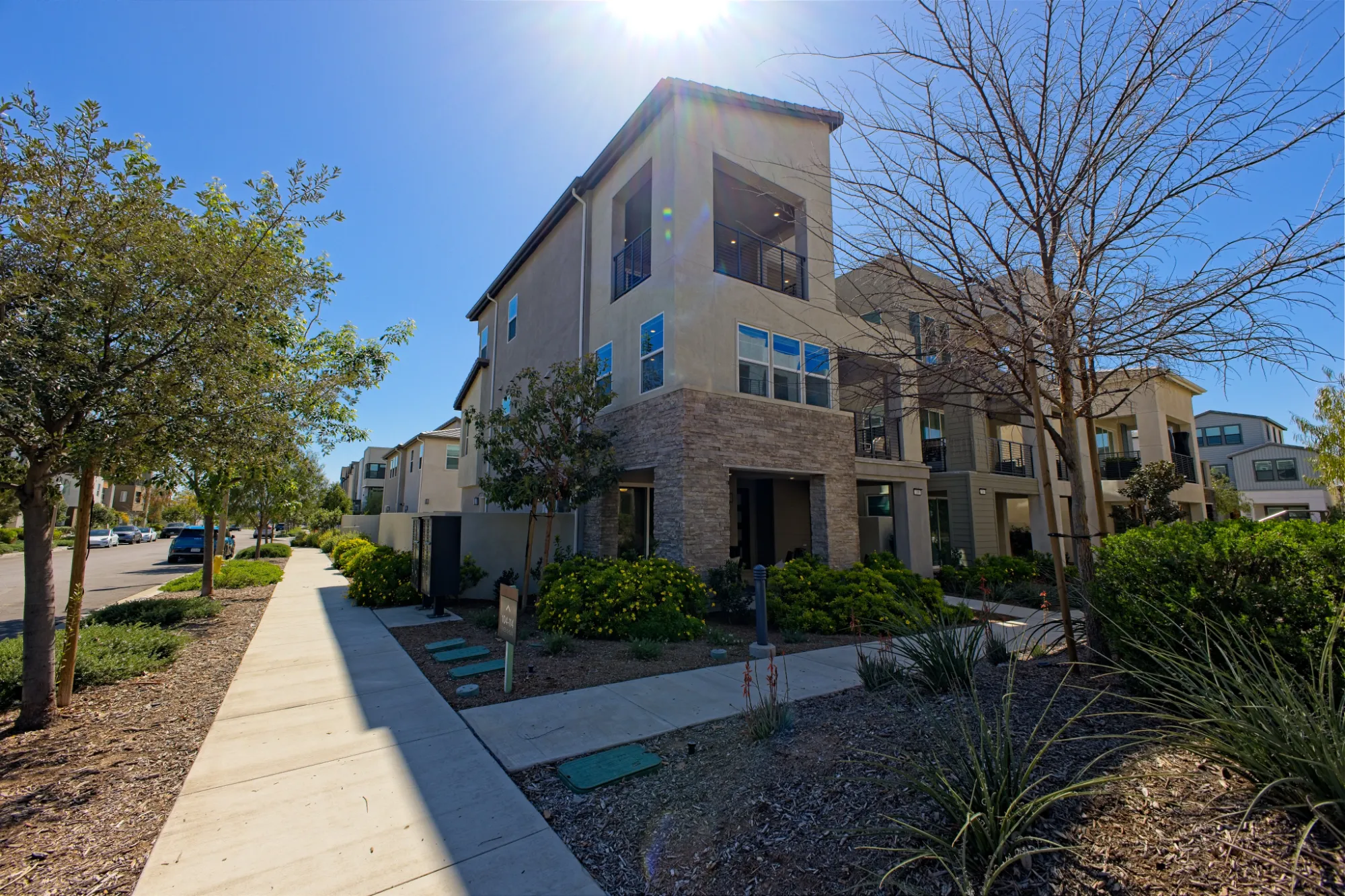 Modern three-story beige apartment building next to a sidewalk with trees and landscaping under a clear blue sky.