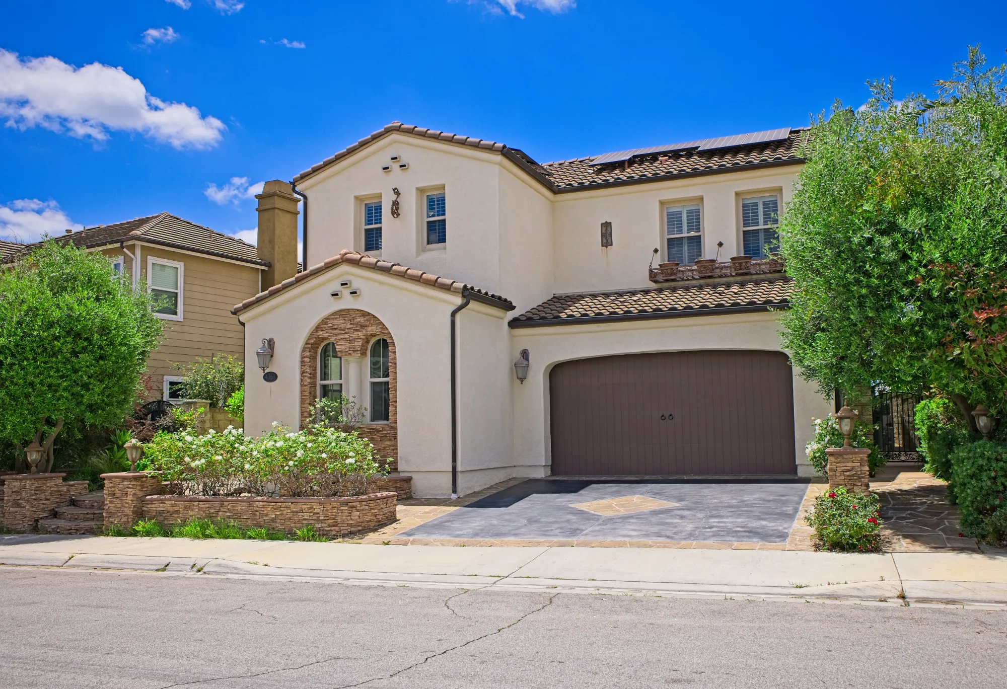 Two-story modern house with a tiled roof, double garage, manicured bushes, and solar panels on roof.