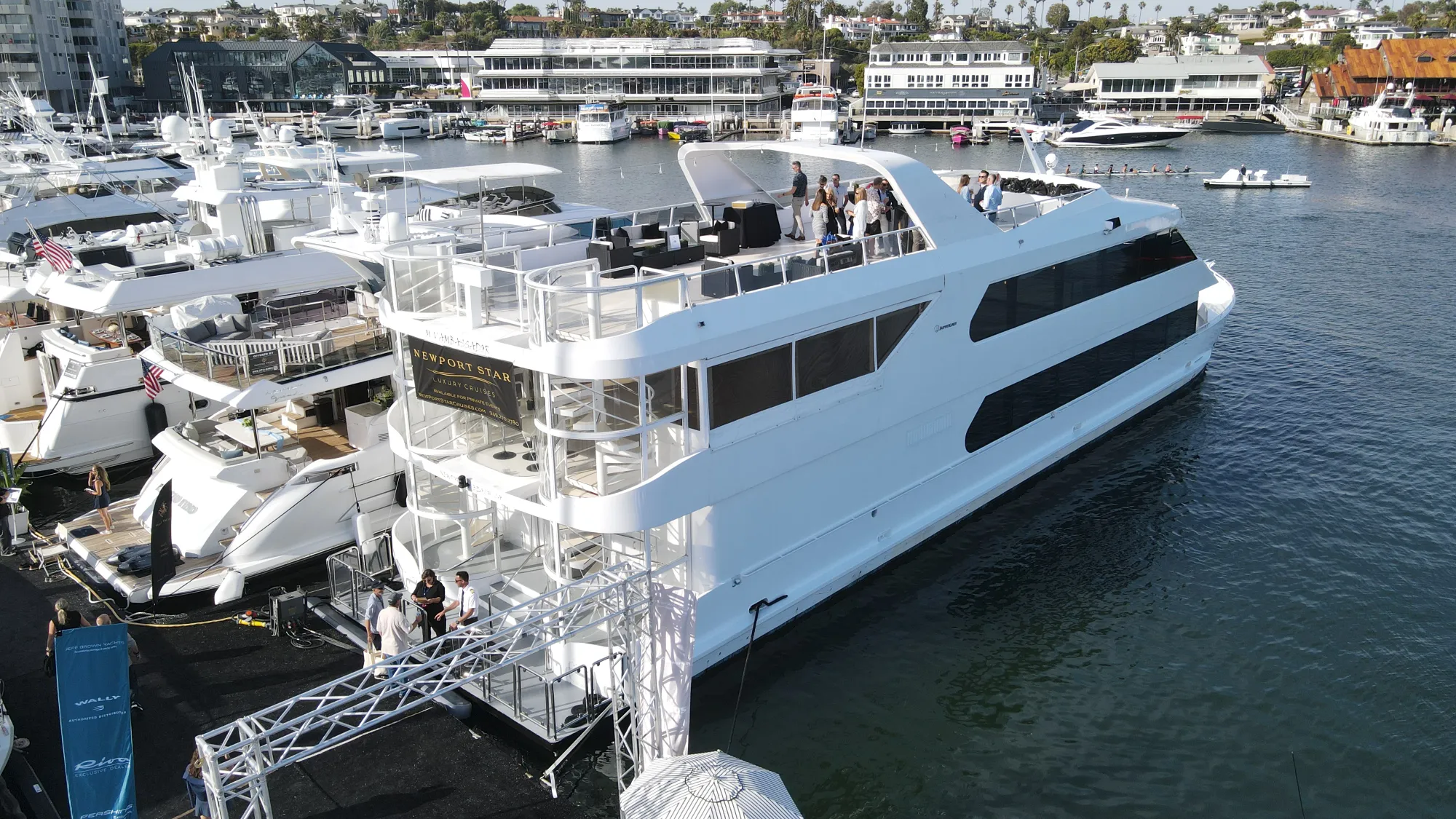 Large white luxury yacht docked at marina with people on upper deck and nearby boats.
