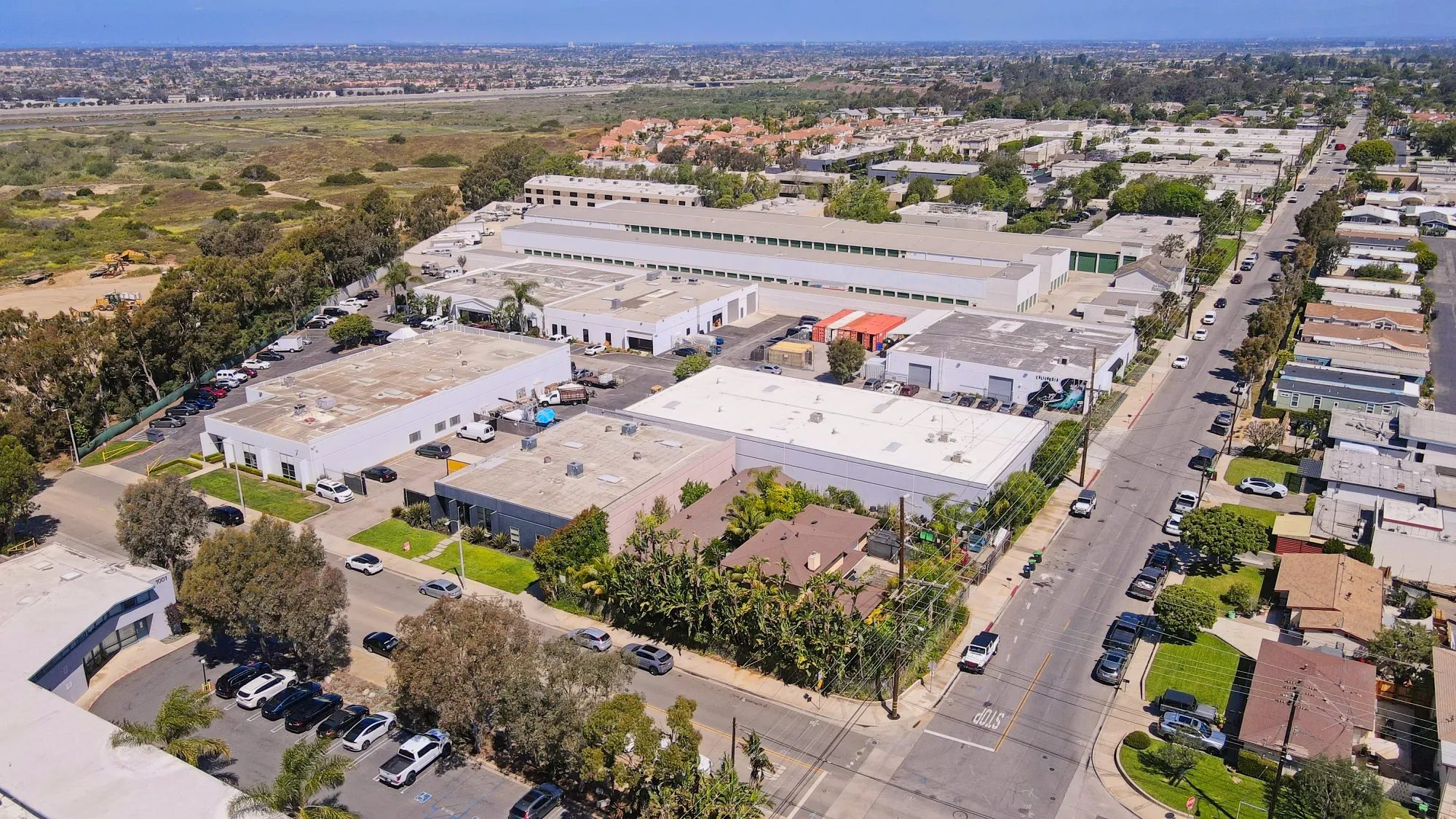 Aerial view of industrial buildings, parking lots, streets, and residential houses under clear sky.