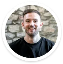 Smiling man with short brown hair and beard wearing a black shirt in front of a stone wall.