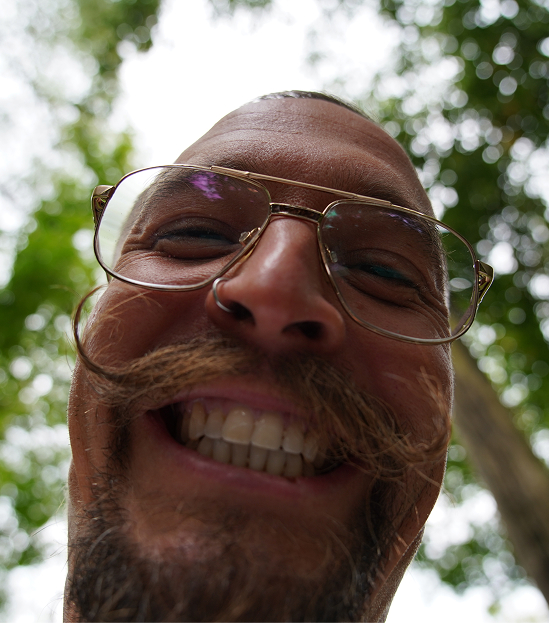 Nahaufnahme eines lachenden Mitarbeiters mit Schnurrbart und Brille im Grünen.