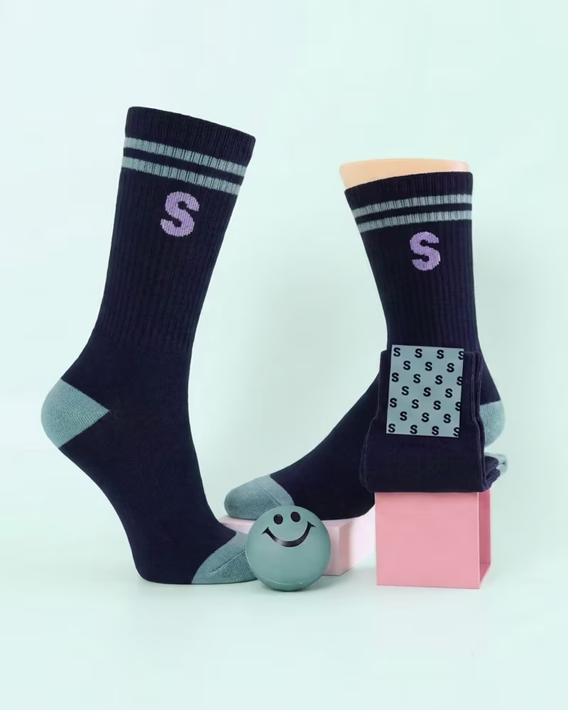 Pair of black socks with gray toe and heel accents, featuring two gray stripes and a purple 'S' near the top, displayed on a mannequin foot and a pink block, with a gray smiley face ball placed in front.