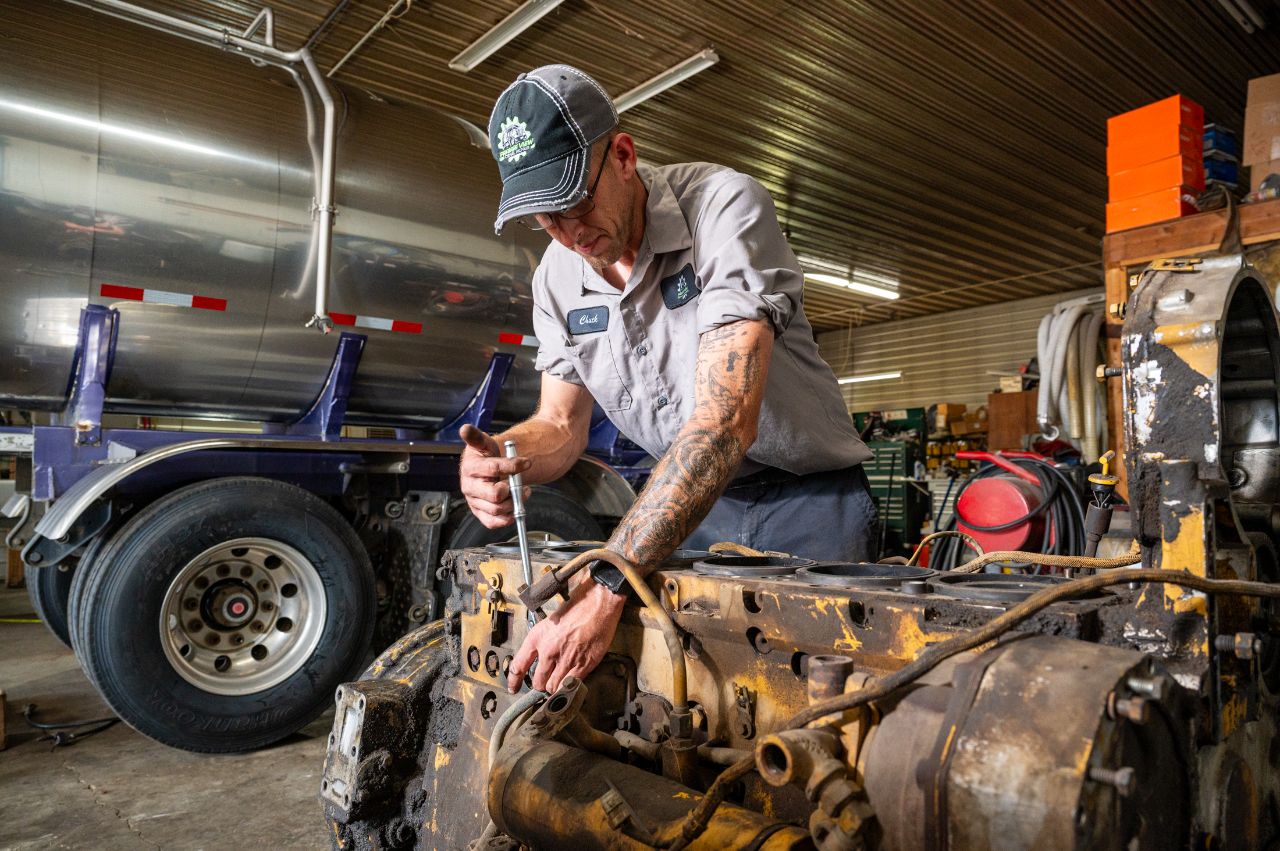 Mechanic working on disassembled diesel engine with wrench inside repair shop, semi-truck trailer in background, surrounded by tools and workshop equipment. 
