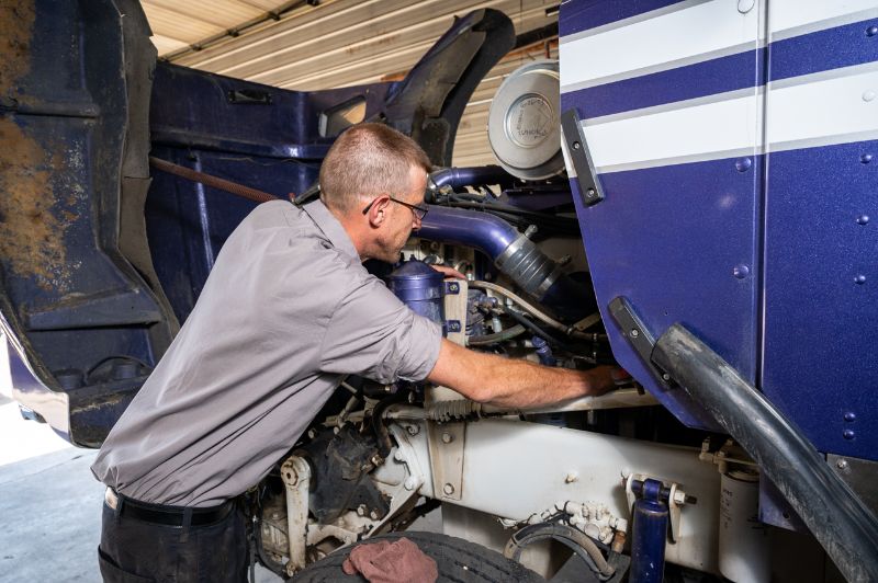 Diesel mechanic working under hood of blue semi-truck inside repair shop, inspecting and repairing engine components with tools, focused on maintenance.