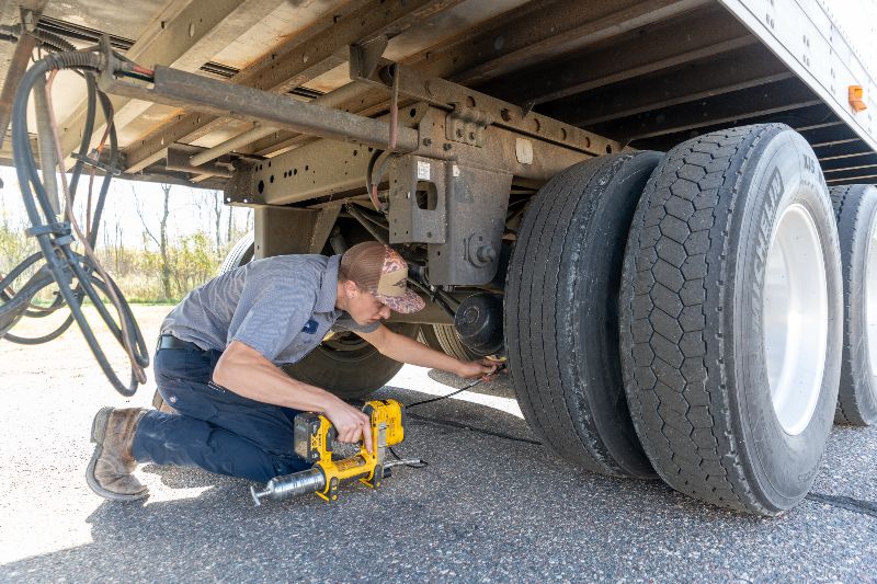Technician kneeling beside a trailer, using tools to service suspension components and check wheels during mobile roadside heavy-duty truck maintenance.
