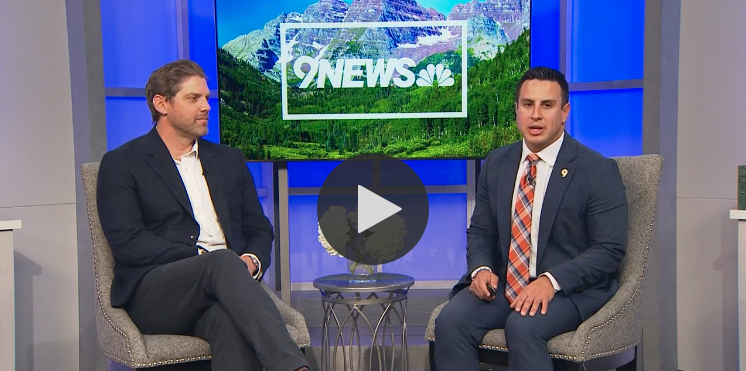 Two men in business attire seated on a talk show set with OneNews logo and mountain backdrop.