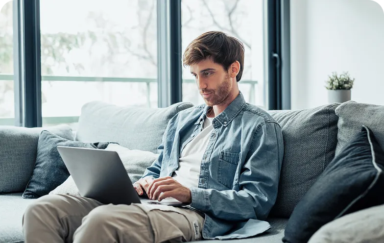 Man sitting on a grey couch working remotely using a laptop near large windows and a small potted plant.