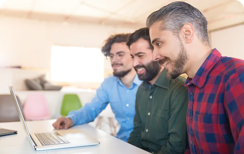 Three men sitting at a table in an office, smiling while looking at a laptop screen together.