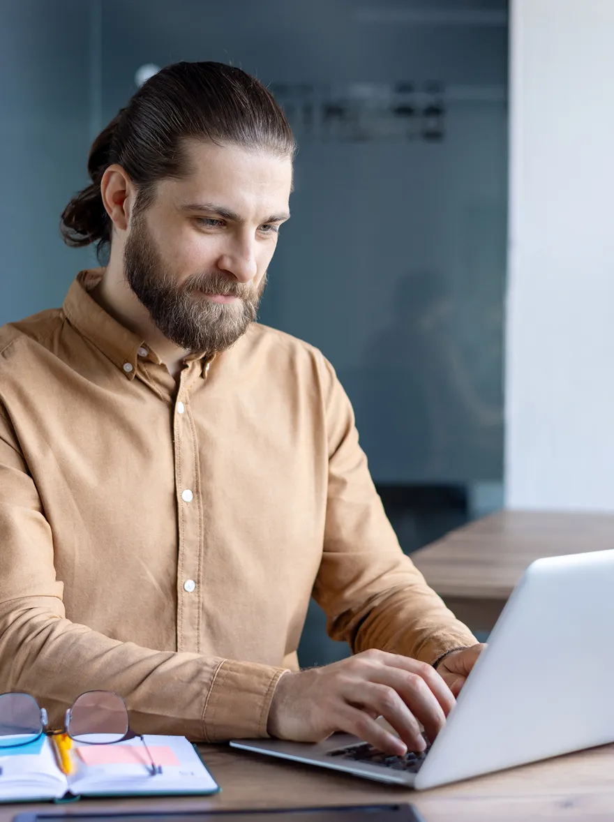 Man in brown shirt typing on a laptop at a desk with glasses and a notebook.