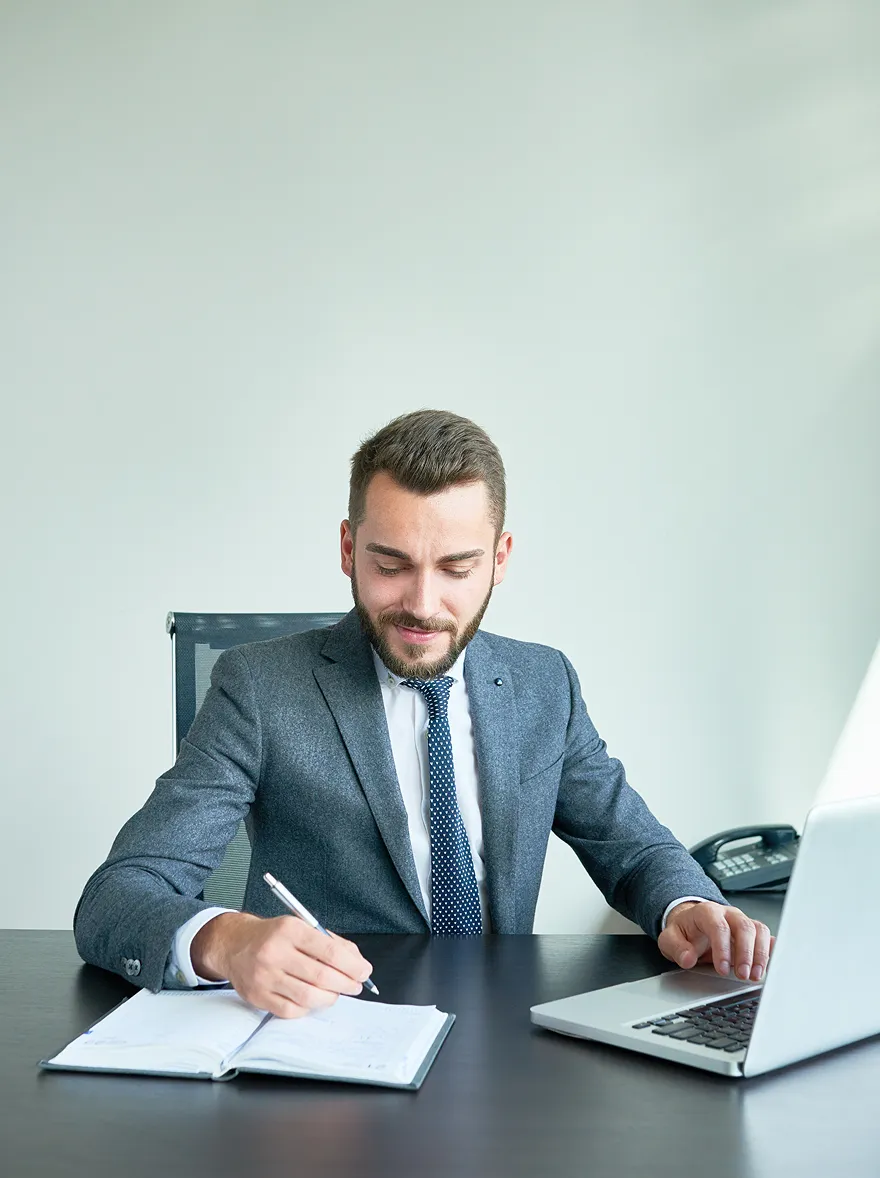 Business professional in a suit writing notes in a notebook while working on a laptop at a modern office desk.
