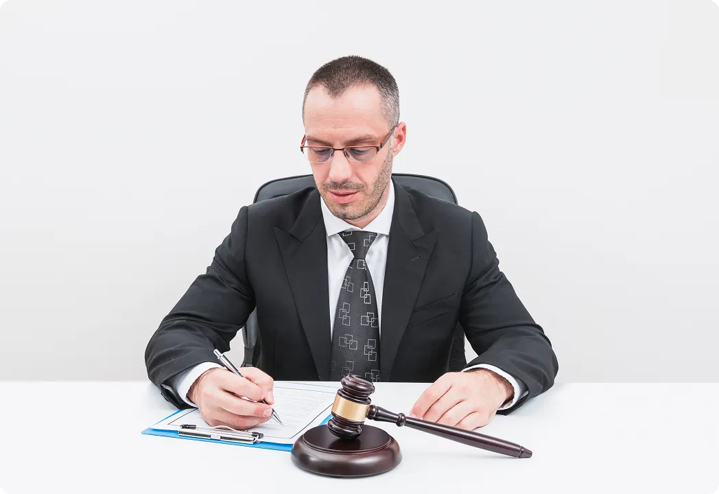 An attorney in a suit writing on a clipboard with a judge's gavel on the desk in front of him.