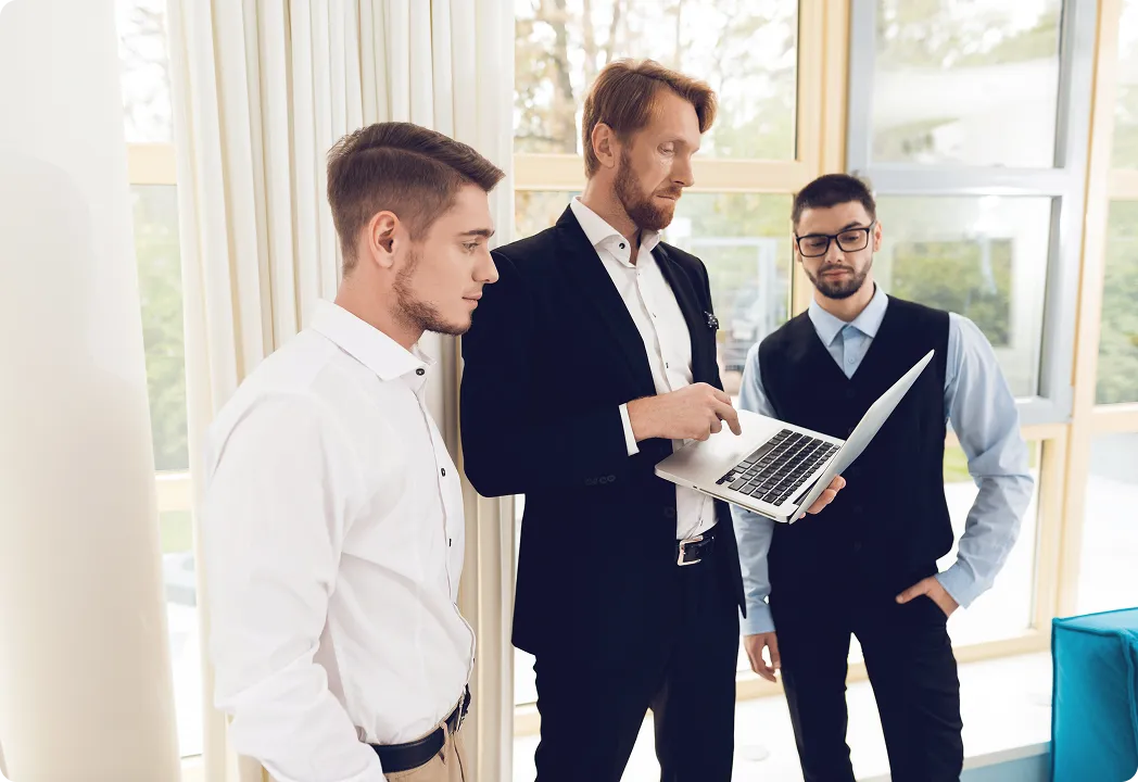 Three men in formal attire standing and discussing something on a laptop.
