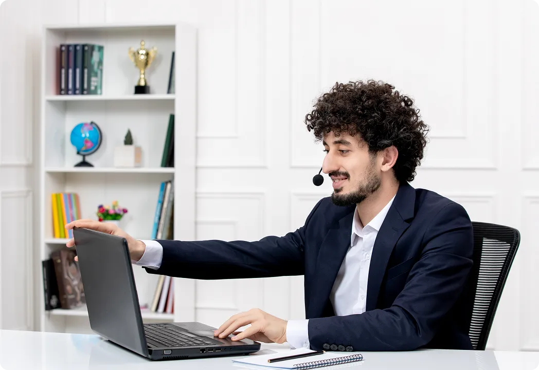 Virtual assistant in a headset and suit smiling while using a laptop in a bright office setting.