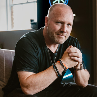 Confident man with a shaved head wearing casual black attire, seated indoors with hands clasped and a thoughtful expression.
