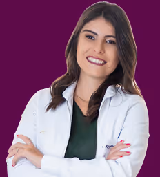 Smiling woman with long dark hair wearing a white lab coat and green top, arms crossed, on a purple background.