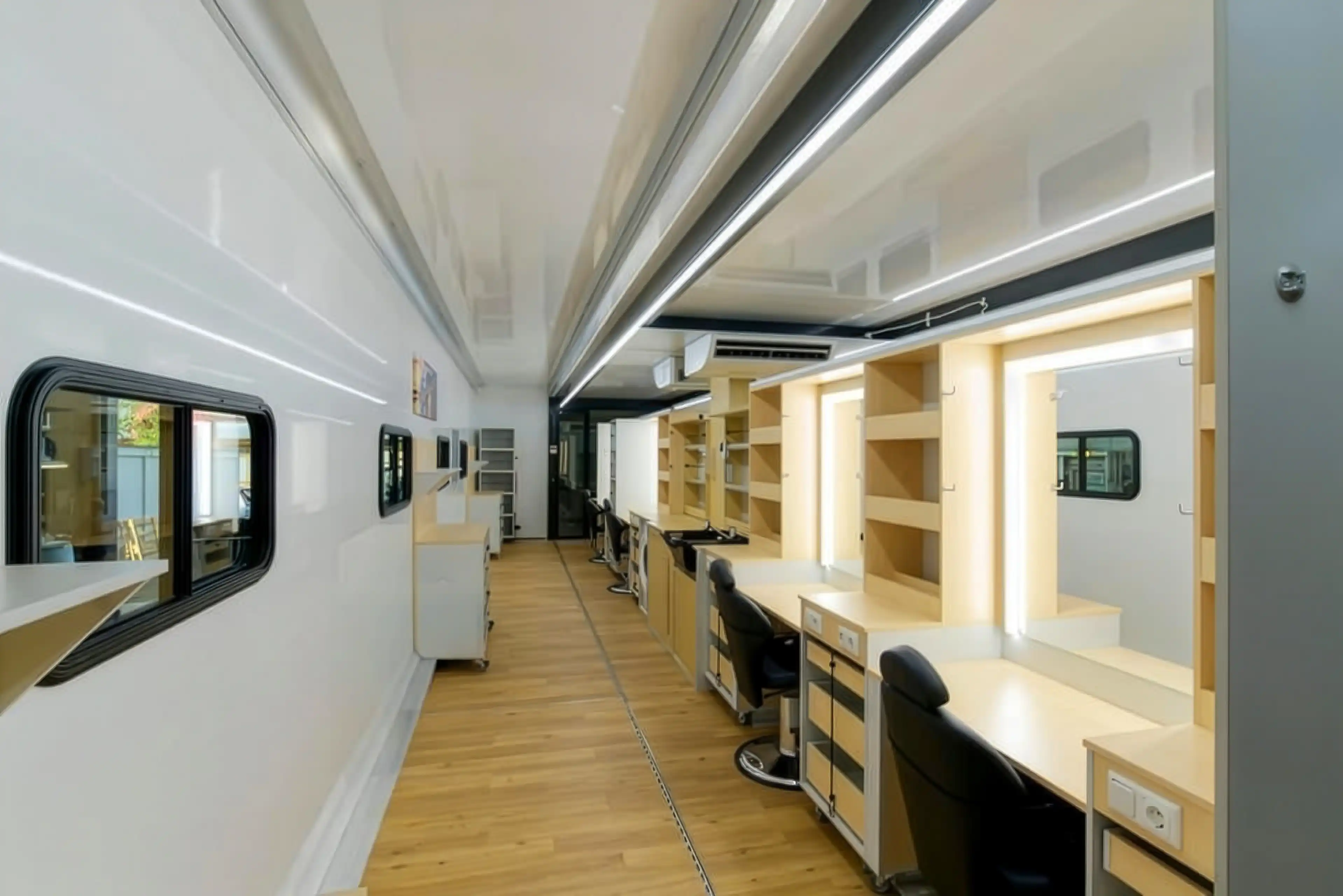 L'intérieur d'une caravane de coiffure et de maquillage avec un long comptoir blanc et des chaises de bureau noires, sous des plafonniers lumineux, prête pour une journée de coiffage et de préparation.
