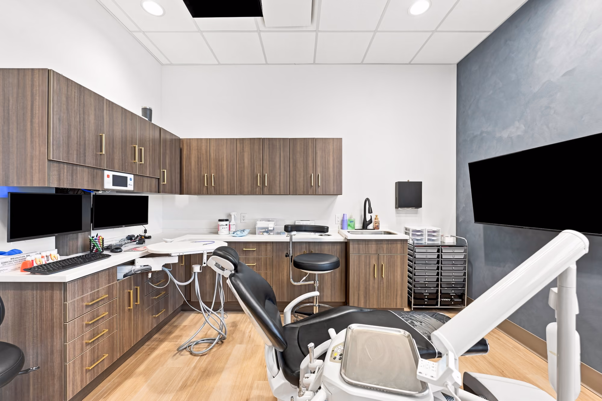 Modern dental exam room with black dental chair, wooden cabinets, computers, and a sink.