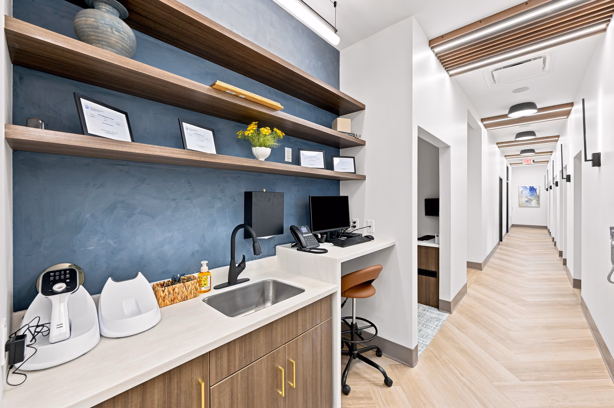 Modern dental office workspace with sink, computer, certificates, and a long corridor with wooden ceiling accents.