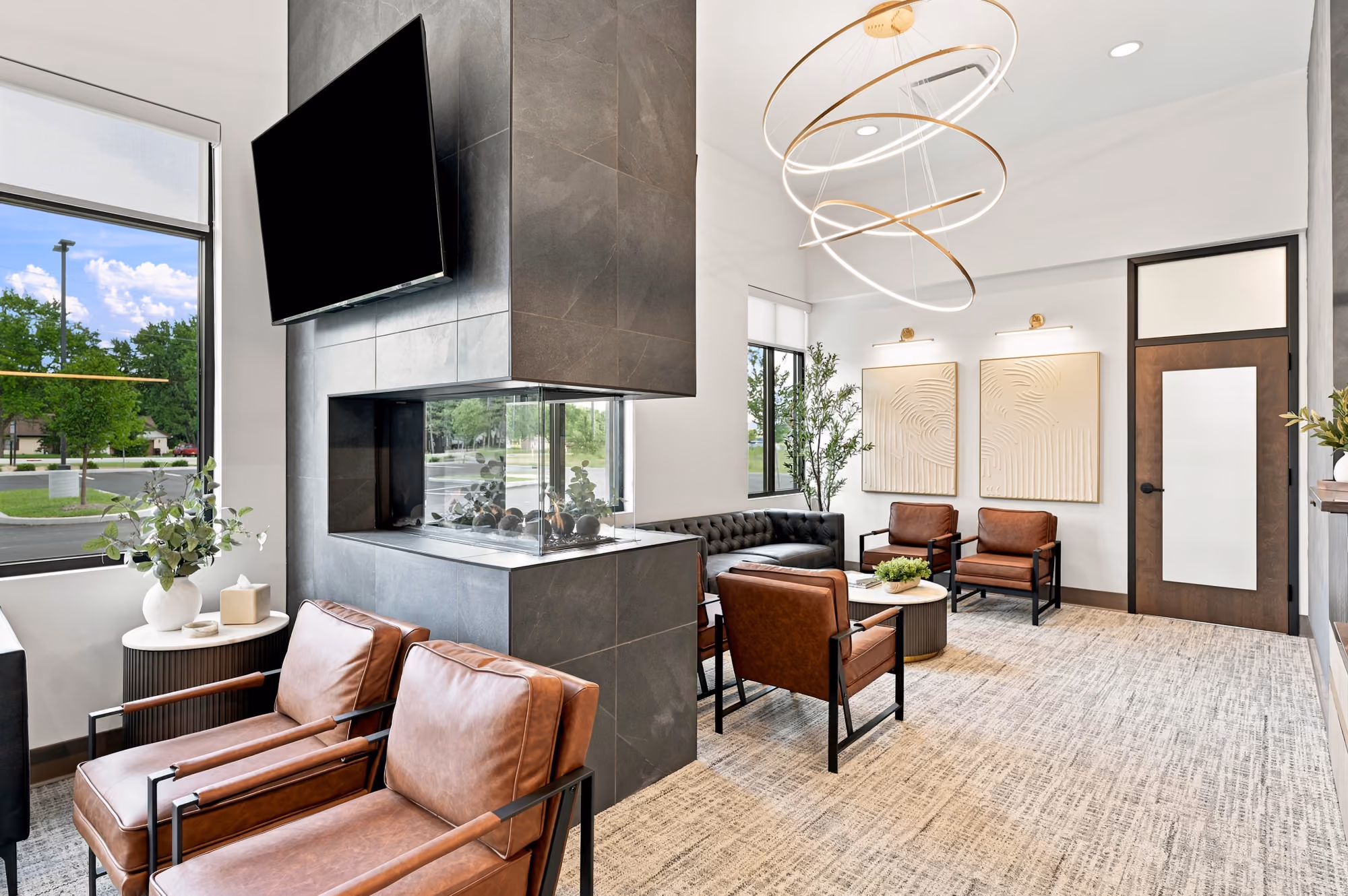 Modern waiting room with leather chairs, gray couch, fireplace, TV, and large windows.