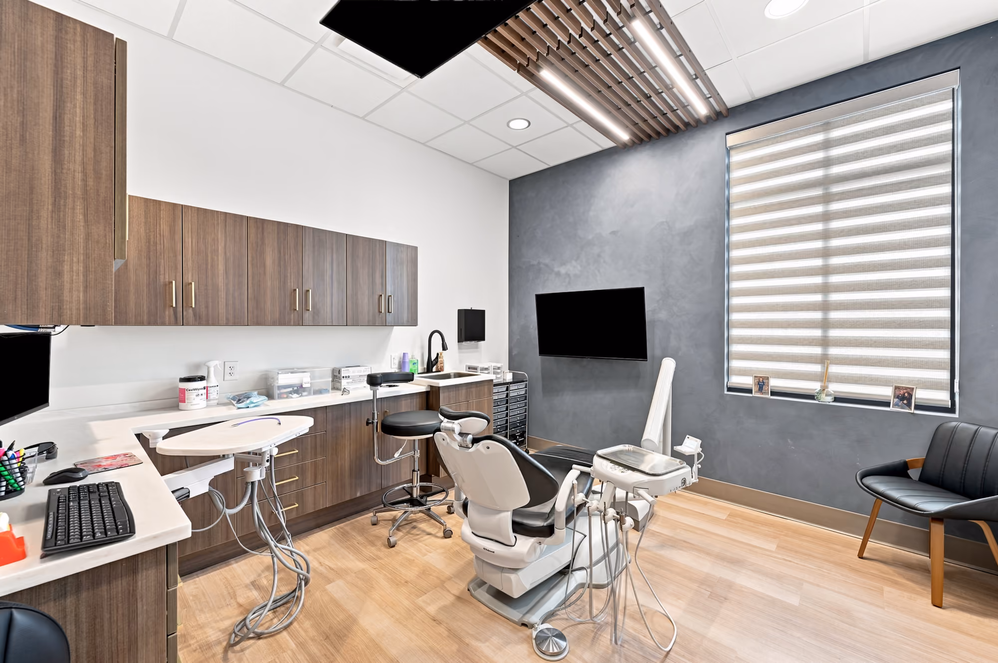 Modern dental exam room with chair, monitors, cabinetry, stool, and window with blinds.