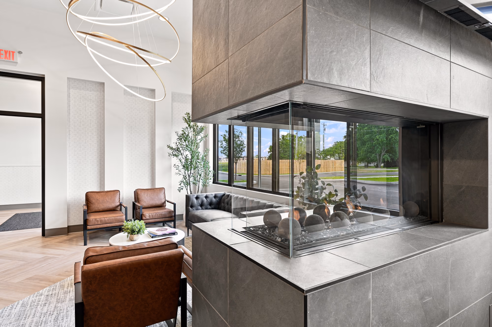 Modern lounge with brown chairs, gray sofa, indoor plants, round light fixture, and corner glass fireplace.