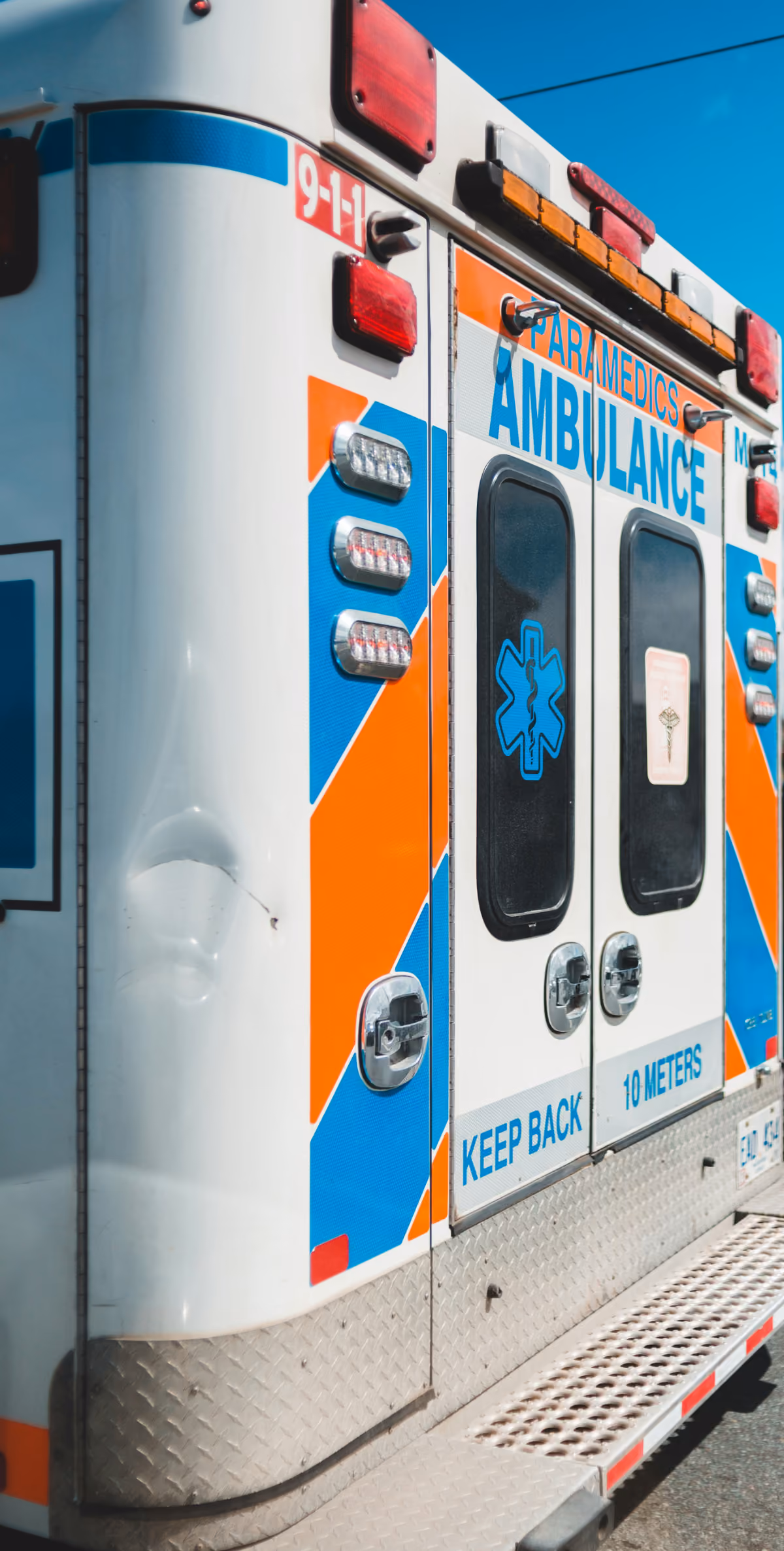 The back of a white ambulance with blue and orange diagonal stripes displays emergency lights, the Star of Life medical symbol, and the warning text "KEEP BACK 10 METERS." A dent is visible on the left rear panel, and the number "9-1-1" is marked near the top.