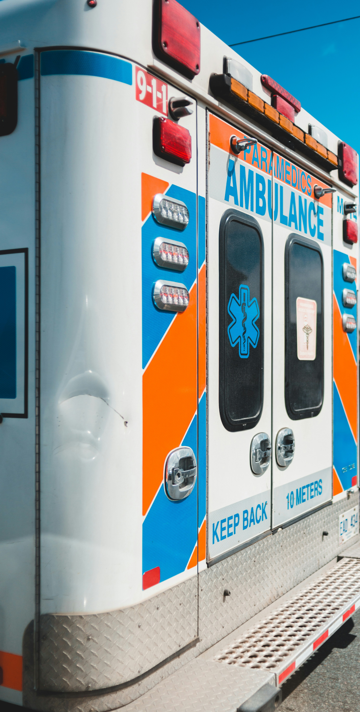 The back of a white ambulance with blue and orange diagonal stripes displays emergency lights, the Star of Life medical symbol, and the warning text "KEEP BACK 10 METERS." A dent is visible on the left rear panel, and the number "9-1-1" is marked near the top.