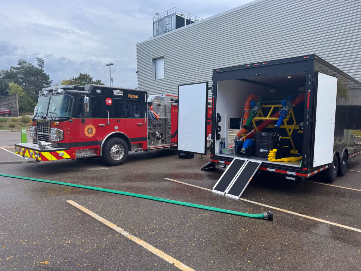 A red fire engine marked “FE2307” and labeled with “ULTIMATE DECON” is parked next to an open black trailer displaying colorful decontamination equipment and hoses. A green hose runs along the wet pavement in a parking lot outside a modern building.