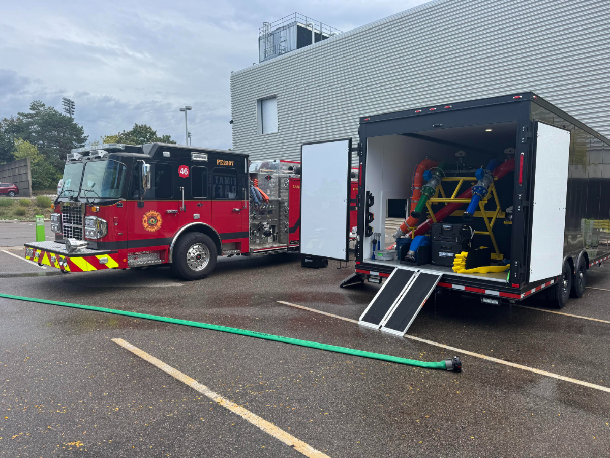 A red fire engine marked “FE2307” and labeled with “ULTIMATE DECON” is parked next to an open black trailer displaying colorful decontamination equipment and hoses. A green hose runs along the wet pavement in a parking lot outside a modern building.