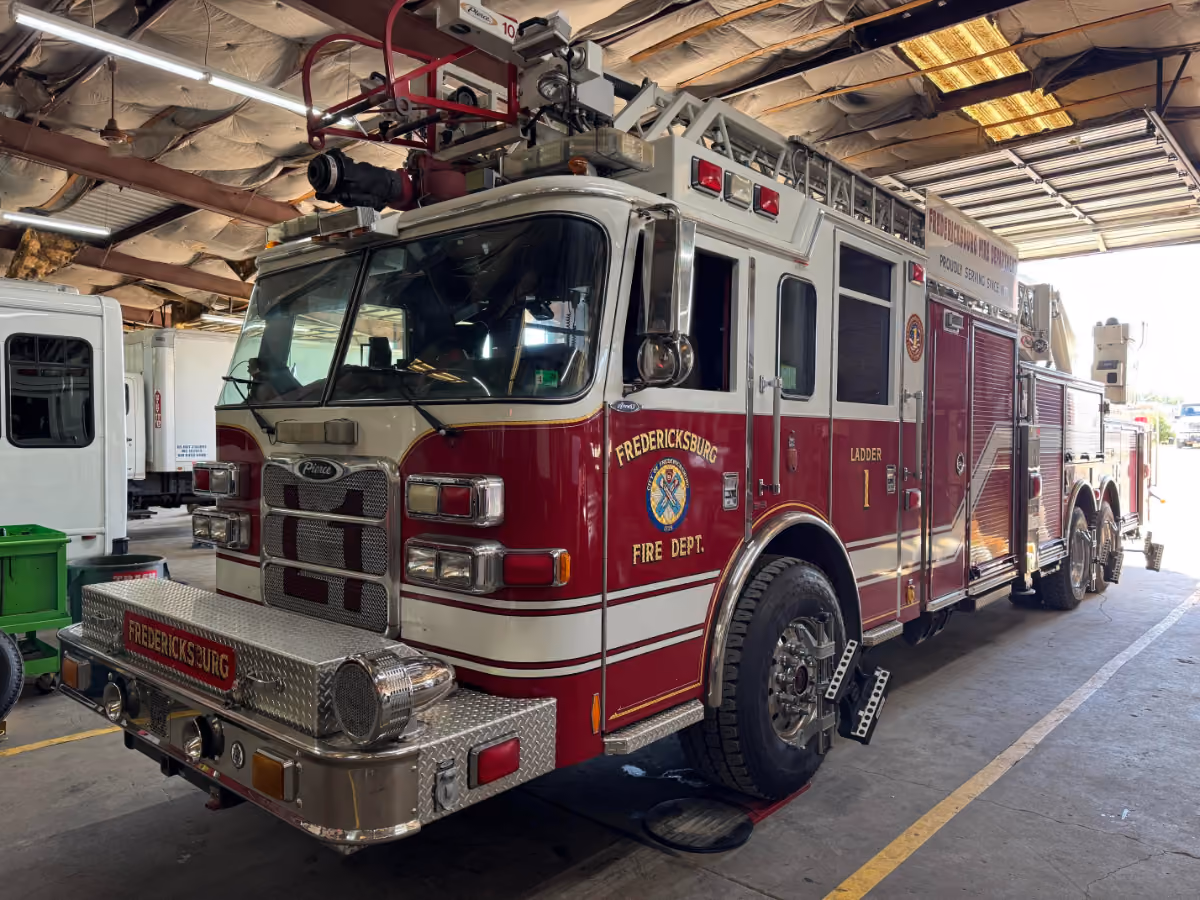 A red Fredericksburg Fire Department Ladder 1 truck is parked inside a service garage with its front wheels secured by metal chocks. The vehicle has a mounted aerial ladder, various compartments, and reflective lettering indicating its department and unit.