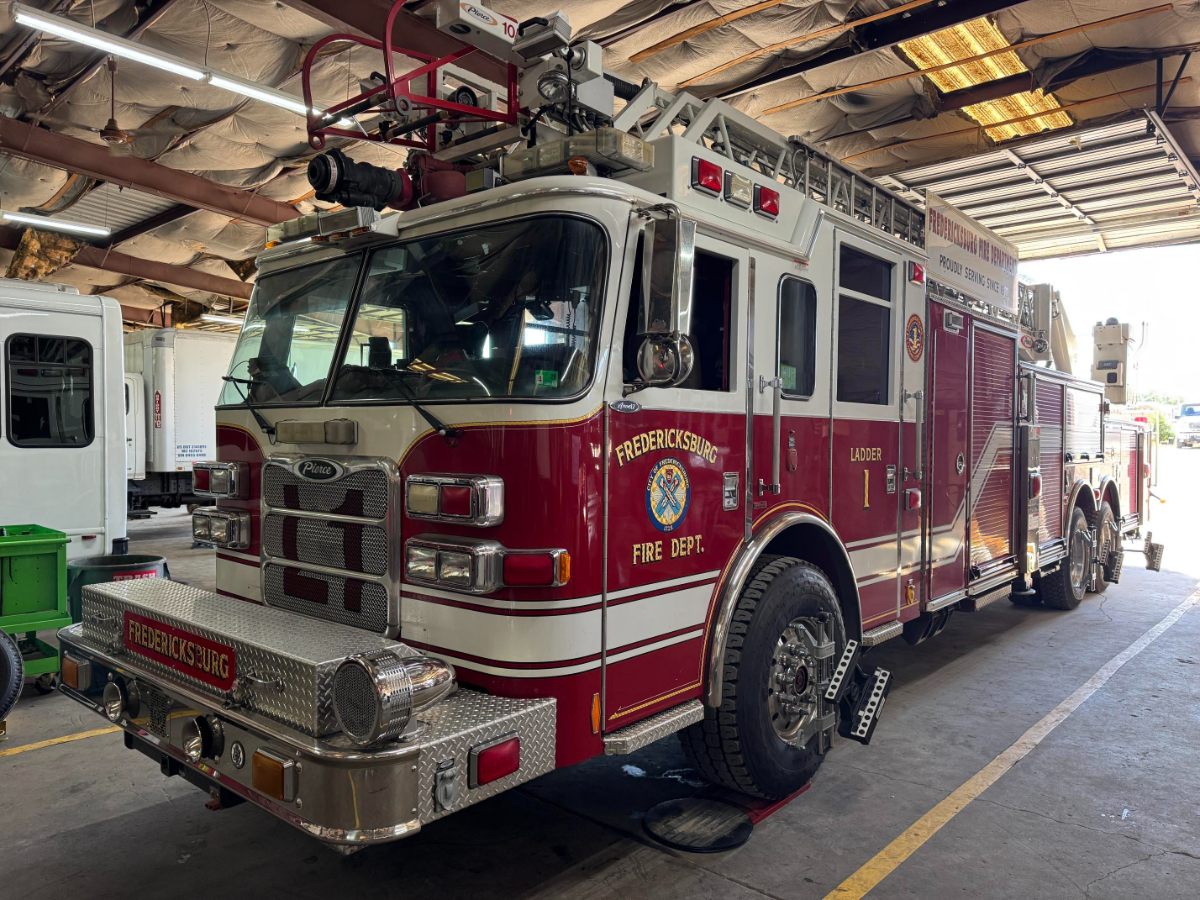 A red Fredericksburg Fire Department Ladder 1 truck is parked inside a service garage with its front wheels secured by metal chocks. The vehicle has a mounted aerial ladder, various compartments, and reflective lettering indicating its department and unit.